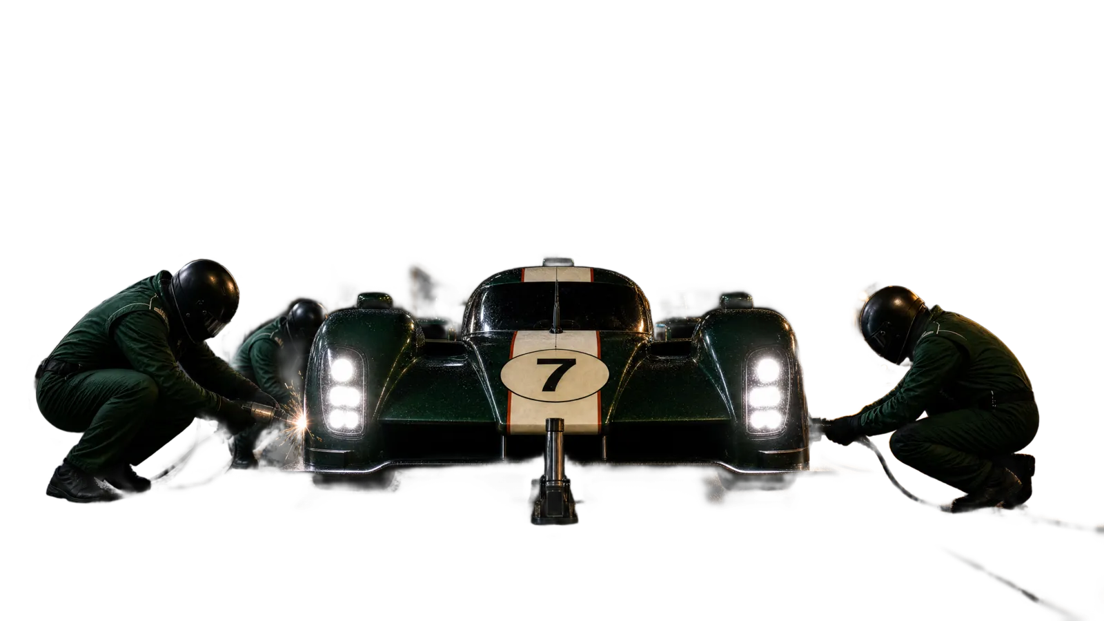 A green endurance race car stopped in its pit box at night, headlights on, while three crew change the front wheels.